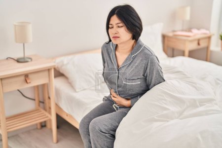 Foto de Joven mujer china que sufre de dolor menstrual sentada en la cama en el dormitorio - Imagen libre de derechos