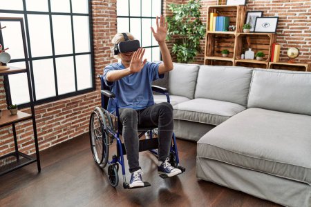 Photo for Young blond man playing video game using virtual reality glasses sitting on wheelchair at home - Royalty Free Image