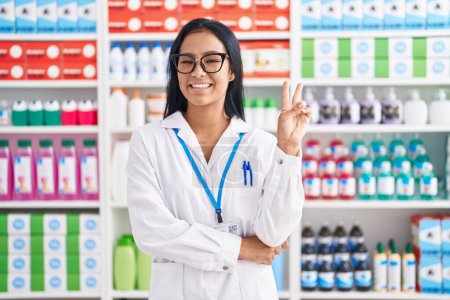 Téléchargez les photos : Femme hispanique travaillant à la pharmacie pharmacie souriant avec un sourire heureux clin d'oeil à la caméra faisant signe de victoire. numéro deux. - en image libre de droit