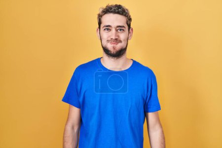 Téléchargez les photos : Hispanic man with beard standing over yellow background puffing cheeks with funny face. mouth inflated with air, crazy expression. - en image libre de droit
