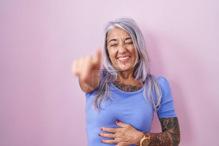 Téléchargez les photos : Femme d'âge moyen avec des tatouages debout sur fond rose se moquant de vous, pointant du doigt l'appareil photo avec la main sur le corps, expression de honte - en image libre de droit