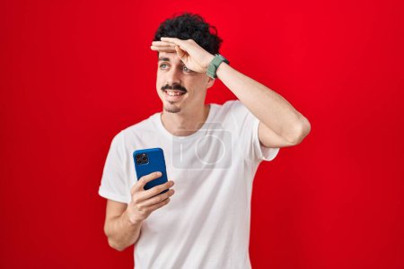 Photo for Hispanic man using smartphone over red background very happy and smiling looking far away with hand over head. searching concept. - Royalty Free Image