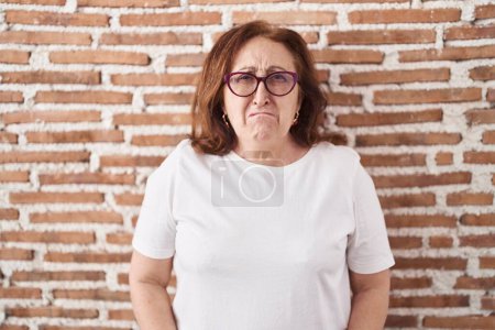 Foto de Mujer mayor con gafas de pie sobre ladrillos pared deprimida y preocuparse por la angustia, llorando enojado y miedo. expresión triste. - Imagen libre de derechos