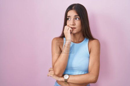 Foto de Young brunette woman standing over pink background looking stressed and nervous with hands on mouth biting nails. anxiety problem. - Imagen libre de derechos