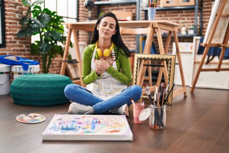 Téléchargez les photos : Femme hispanique assise au studio d'art peignant sur toile souriant avec les mains sur la poitrine avec les yeux fermés et geste reconnaissant sur le visage. concept de santé. - en image libre de droit