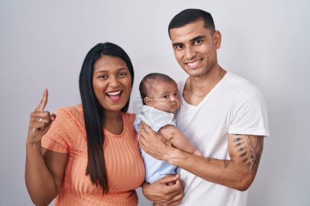 Photo for Young hispanic couple with baby standing together over isolated background smiling amazed and surprised and pointing up with fingers and raised arms. - Royalty Free Image