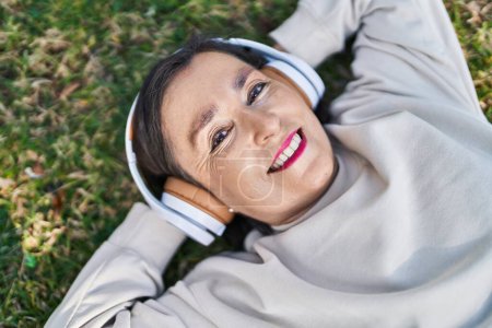 Photo for Middle age woman listening to music lying on herb at park - Royalty Free Image