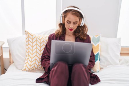 Foto de Young beautiful hispanic woman using laptop and headphones sitting on bed at bedroom - Imagen libre de derechos