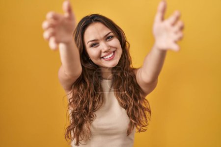 Photo for Young hispanic woman standing over yellow background looking at the camera smiling with open arms for hug. cheerful expression embracing happiness. - Royalty Free Image
