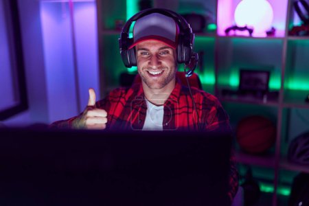 Foto de Joven hombre caucásico jugando videojuegos sonriendo feliz y positivo, pulgar hacia arriba haciendo excelente y signo de aprobación - Imagen libre de derechos