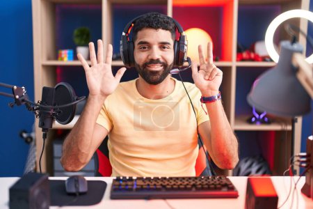 Foto de Hombre hispano con barba jugando videojuegos con auriculares mostrando y apuntando hacia arriba con los dedos número ocho mientras sonríe confiado y feliz. - Imagen libre de derechos