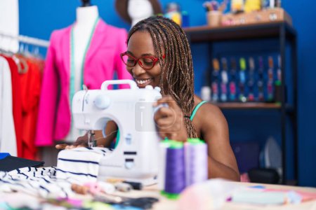 Téléchargez les photos : Femme afro-américaine tailleur souriant confiant en utilisant une machine à coudre à l'atelier - en image libre de droit