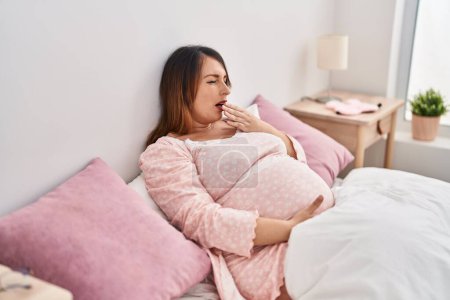 Téléchargez les photos : Jeune femme enceinte bâillant assis sur le lit dans la chambre - en image libre de droit