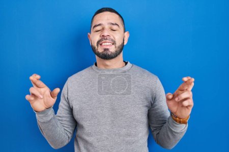 Photo for Hispanic man standing over blue background gesturing finger crossed smiling with hope and eyes closed. luck and superstitious concept. - Royalty Free Image