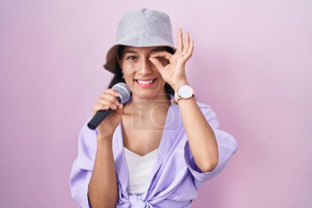 Téléchargez les photos : Jeune femme hispanique chantant chanson à l'aide d'un microphone sur fond rose souriant heureux faire ok signe avec la main sur les yeux en regardant à travers les doigts - en image libre de droit