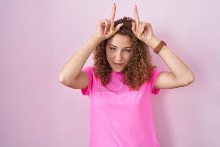 Photo for Young caucasian woman standing over pink background doing funny gesture with finger over head as bull horns - Royalty Free Image