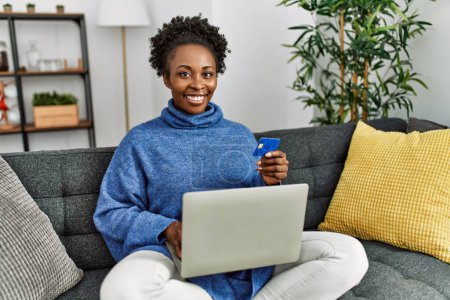 Foto de Mujer afroamericana usando laptop y tarjeta de crédito sentada en sofá en casa - Imagen libre de derechos