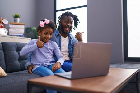 Photo for African father and daughter doing video call on laptop at home pointing thumb up to the side smiling happy with open mouth - Royalty Free Image