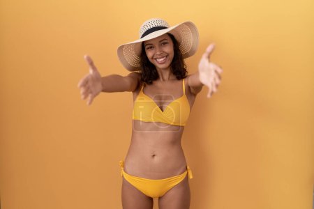Téléchargez les photos : Jeune femme hispanique portant un bikini et un chapeau d'été regardant la caméra souriant à bras ouverts pour un câlin. expression joyeuse embrassant le bonheur. - en image libre de droit