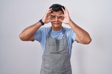 Photo for Hispanic young man wearing apron over white background trying to open eyes with fingers, sleepy and tired for morning fatigue - Royalty Free Image