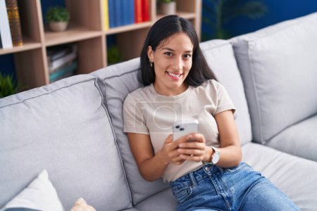 Foto de Mujer hispana hermosa joven usando teléfono inteligente sentado en el sofá en casa - Imagen libre de derechos