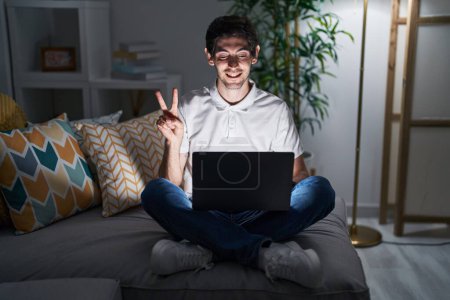Foto de Joven hombre hispano usando portátil en casa por la noche sonriendo con la cara feliz guiñando a la cámara haciendo signo de victoria con los dedos. número dos. - Imagen libre de derechos