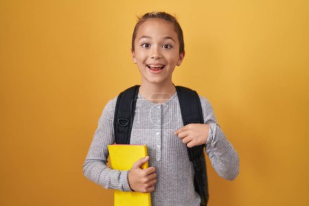 Téléchargez les photos : Petit garçon caucasien portant un sac à dos étudiant et tenant un livre pointant du doigt un sourire heureux et fier - en image libre de droit
