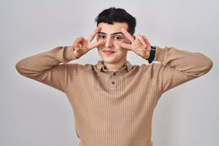 Photo for Non binary person standing over isolated background doing peace symbol with fingers over face, smiling cheerful showing victory - Royalty Free Image