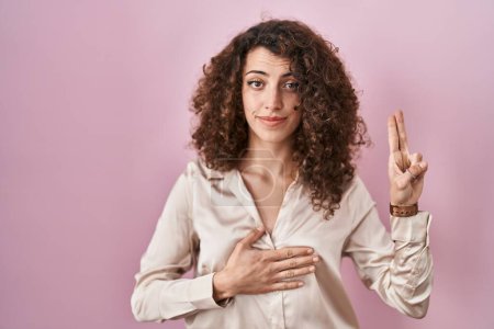 Téléchargez les photos : Femme hispanique avec les cheveux bouclés debout sur fond rose souriant jurant avec la main sur la poitrine et les doigts vers le haut, faisant un serment de fidélité promesse - en image libre de droit