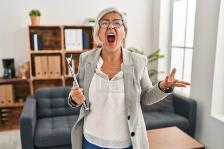 Photo for Middle age woman with grey hair at consultation office angry and mad screaming frustrated and furious, shouting with anger. rage and aggressive concept. - Royalty Free Image