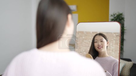 Téléchargez les photos : Young chinese woman combing hair looking on mirror at bedroom - en image libre de droit