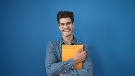 Foto de Young hispanic man smiling confident holding book over isolated blue background - Imagen libre de derechos