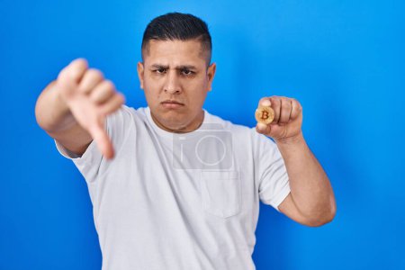 Photo for Hispanic young man holding virtual currency bitcoin with angry face, negative sign showing dislike with thumbs down, rejection concept - Royalty Free Image