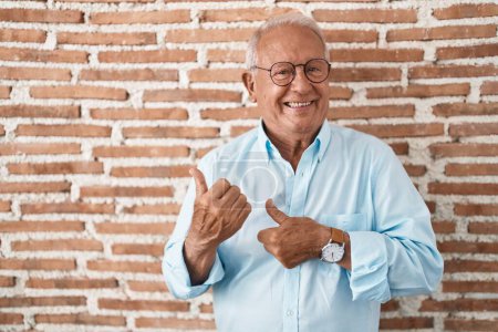 Téléchargez les photos : Homme âgé avec les cheveux gris debout sur le mur de briques pointant vers le dos derrière avec la main et les pouces vers le haut, souriant confiant - en image libre de droit