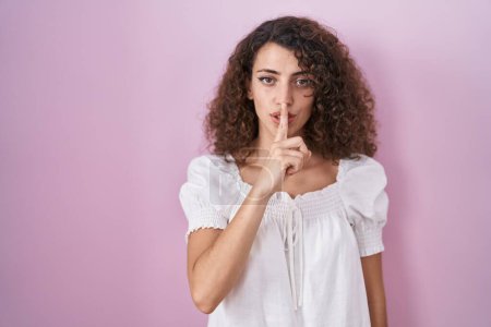 Photo for Hispanic woman with curly hair standing over pink background asking to be quiet with finger on lips. silence and secret concept. - Royalty Free Image