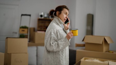 Foto de Mujer hispana de mediana edad hablando en el teléfono inteligente bebiendo café en un nuevo hogar - Imagen libre de derechos