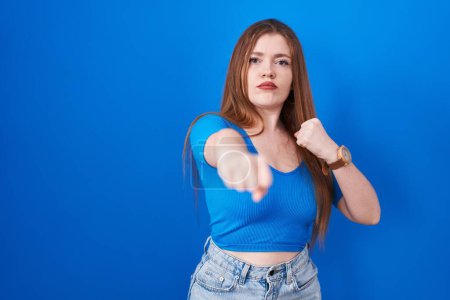 Téléchargez les photos : Femme rousse debout sur fond bleu coup de poing poing pour se battre, attaque agressive et en colère, menace et violence - en image libre de droit