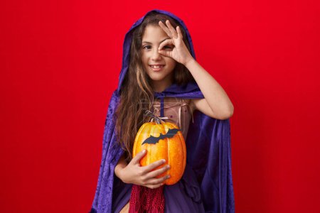 Foto de Pequeña chica hispana vistiendo disfraz de bruja halloween sonriendo feliz haciendo señal de bien con la mano en el ojo mirando a través de los dedos - Imagen libre de derechos