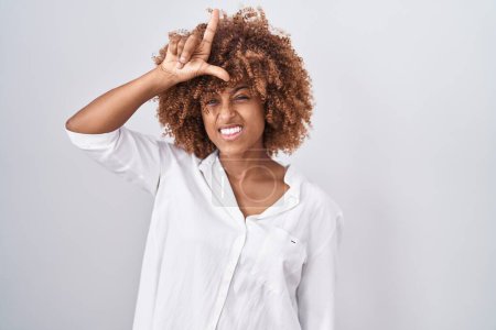 Photo for Young hispanic woman with curly hair standing over white background making fun of people with fingers on forehead doing loser gesture mocking and insulting. - Royalty Free Image