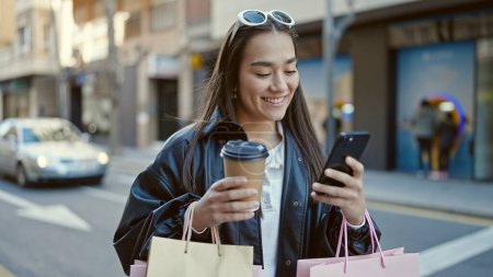 Téléchargez les photos : Jeune belle femme hispanique en utilisant un smartphone tenant des sacs à provisions et du café dans la rue - en image libre de droit