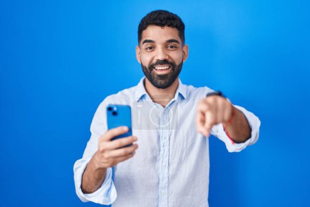 Téléchargez les photos : Homme hispanique avec barbe en utilisant un message de dactylographie smartphone pointant vers vous et la caméra avec les doigts, souriant positif et joyeux - en image libre de droit