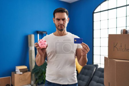 Téléchargez les photos : Young hispanic man with beard holding piggy bank and credit card at new home skeptic and nervous, frowning upset because of problem. negative person. - en image libre de droit