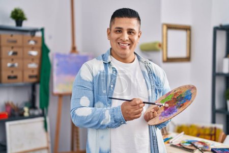 Téléchargez les photos : Jeune homme latino artiste souriant confiant tenant palette et pinceau au studio d'art - en image libre de droit