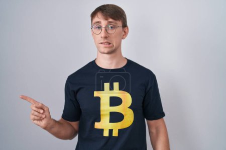 Foto de Hombre rubio caucásico con camiseta bitcoin señalando a un lado preocupado y nervioso con el dedo índice, preocupado y expresión sorprendida - Imagen libre de derechos