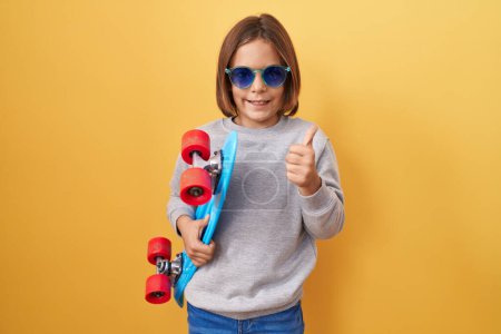 Photo for Little hispanic boy wearing sunglasses holding skate smiling happy and positive, thumb up doing excellent and approval sign - Royalty Free Image
