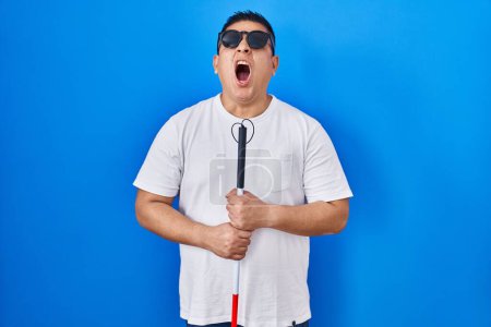 Foto de Joven ciego hispano sosteniendo bastón enojado y loco gritando frustrado y furioso, gritando con ira mirando hacia arriba. - Imagen libre de derechos