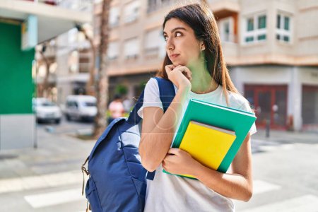 Téléchargez les photos : Jeune belle étudiante hispanique tenant des livres avec une expression de doute dans la rue - en image libre de droit