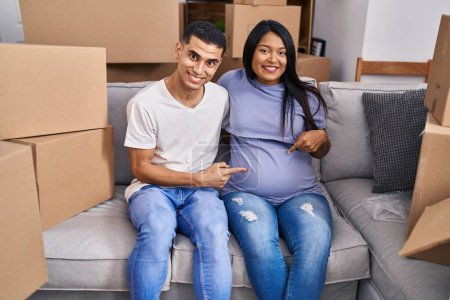 Téléchargez les photos : Jeune couple hispanique s'attendant à un bébé assis sur le canapé à la nouvelle maison souriant heureux pointant avec la main et le doigt - en image libre de droit