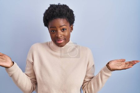 Foto de Mujer afroamericana de pie sobre fondo azul expresión despistada y confusa con los brazos y las manos levantadas. concepto de duda. - Imagen libre de derechos