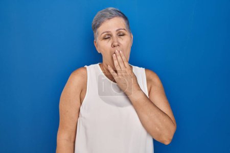 Foto de Mujer caucásica de mediana edad de pie sobre fondo azul aburrido bostezo cansado cubriendo la boca con la mano. inquietud y somnolencia. - Imagen libre de derechos
